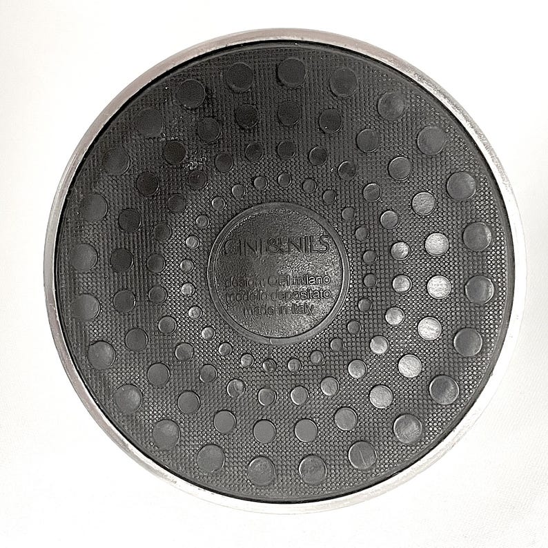 May include: A circular black rubber base with a metallic silver rim. The center features embossed text reading CINI&NILS, design OPI milano, modello depositato, and made in italy. The surface has a textured pattern of raised black circles.