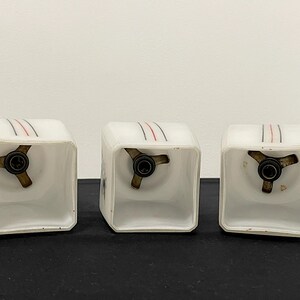 May include: Three vintage white glass light fixtures with a square shape. Each fixture has a brass-coloured base and is decorated with three parallel lines in red and black. The fixtures are displayed on a black surface.