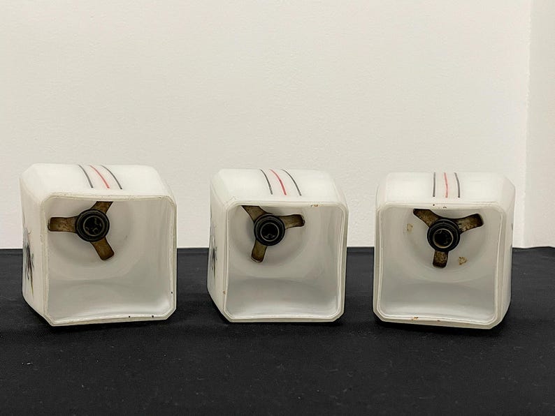 May include: Three vintage white glass light fixtures with a square shape. Each fixture has a brass-coloured base and is decorated with three parallel lines in red and black. The fixtures are displayed on a black surface.