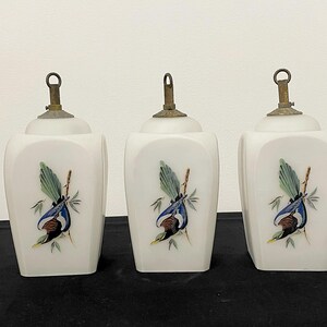 May include: Three white ceramic jars with brass-coloured lids. Each jar features a hand-painted bird design with blue, green, and brown colours. The jars have a square shape with rounded edges.