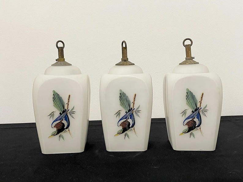 May include: Three white ceramic jars with brass-coloured lids. Each jar features a hand-painted bird design with blue, green, and brown colours. The jars have a square shape with rounded edges.