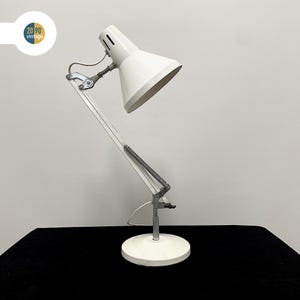 Op de afbeelding: Een witte vintage bureaulamp met een verstelbare arm en een conische kap. De lamp heeft een ronde voet en een metalen frame. De lamp staat op een zwart oppervlak. De lamp heeft een klassiek ontwerp.