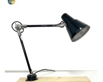 Peut inclure: Lampe de bureau noire avec un abat-jour conique et un bras réglable. La lampe est montée sur une base en bois. La lampe a une esthétique vintage et convient à un bureau ou un espace de travail.
