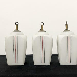 May include: Three vintage white glass pendant lights with brass-coloured hardware. Each light has a vertical design of a black and red stripe. The lights are on a black surface.