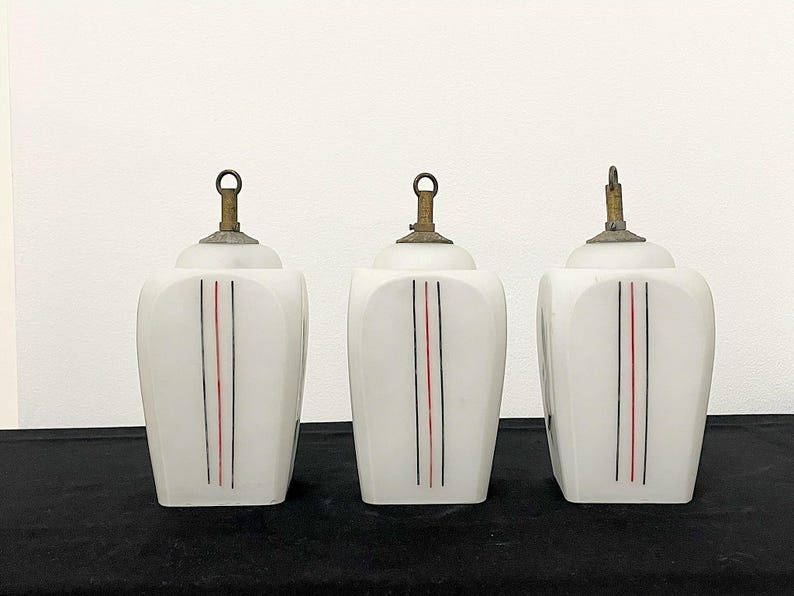 May include: Three vintage white glass pendant lights with brass-coloured hardware. Each light has a vertical design of a black and red stripe. The lights are on a black surface.
