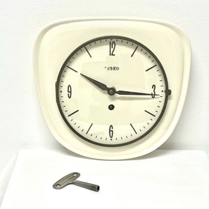 May include: A white ceramic clock with black hands and black Roman numeral markings. The clock face has the word "TESEO" printed on it. A small, silver key is included.