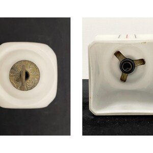 May include: Two views of a white, square-shaped light fitting. One view shows the top with a brass-coloured decorative element. The other view shows the bottom with a three-pronged metal piece and a light bulb socket.