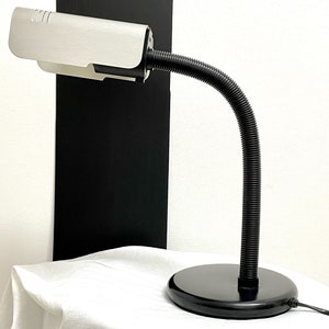 以下が含まれることがあります： 黒と銀のデスクランプ。フレキシブルなグースネックアームと丸いベースが付いています。ランプには、銀色の仕上げが施された白いシェードが付いています。