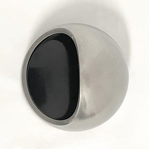 May include: A spherical object with a smooth, metallic silver exterior and a large, circular, matte black inset on one side. The object has a curved, bowl-like indentation that creates a distinct contrast between the reflective silver and the dark center.