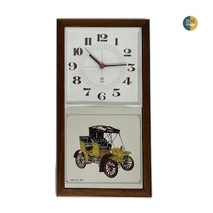 May include: Vintage rectangular wall clock with a brown wooden frame. The top section features a white clock face with black numerals and red hands. The bottom section displays a yellow antique car illustration. The text "CADILLAC 1906" is visible.