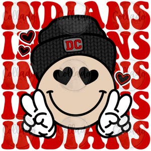 以下が含まれることがあります： ハート型の目とピースサインのスマイリーフェイスが特徴のグラフィックデザイン。スマイリーフェイスは「DC」パッチが付いた黒いビーニー帽をかぶっています。赤いハートと「INDIANS」の文字もデザインに含まれています。