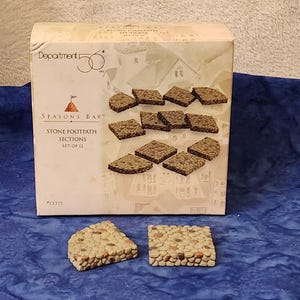 May include: A box of Seasons Bay Stone Footpath Sections, set of 12. The box features a picture of the stone sections. Two stone sections are in front of the box. The stone sections are made of small, tan and brown stones.