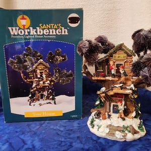 May include: A porcelain lighted house accessory from Santa's Workbench, featuring a tree house design. The miniature house has a snow-covered base, with figures and a sign that reads "Elf's Inn & Shop". The box reads "Tree House".