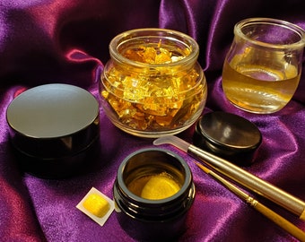 May include: An assortment of beauty products, including small jars and containers. One jar is filled with gold leaf flakes. Other items include a small jar with liquid, a small container with a gold substance, and makeup brushes. The items are arranged on a purple satin surface.