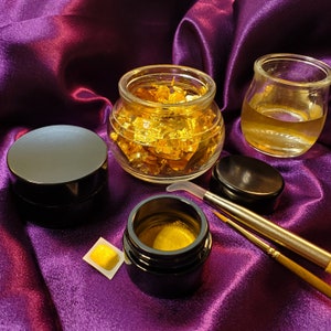Könnte beinhalten: Ein Satz von vier goldfarbenen Gläsern mit Deckeln, von denen zwei mit Goldblättchen gefüllt sind. Die Gläser stehen auf einer violetten Stoffoberfläche. Es befinden sich zwei Pinsel und ein kleines Quadrat aus Blattgold auf der Oberfläche.
