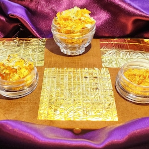 May include: Three glass jars filled with gold leaf flakes on a brown surface with gold leaf squares. The jars are on a purple fabric background.