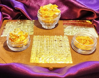May include: Three glass jars filled with gold leaf flakes on a brown surface with gold leaf squares. The jars are on a purple fabric background.