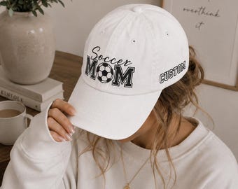 May include: A white baseball cap featuring the black text Soccer MOM with a soccer ball graphic replacing the letter O. The word CUSTOM is printed in black on the side of the cap. The cap has a structured crown and a curved brim.