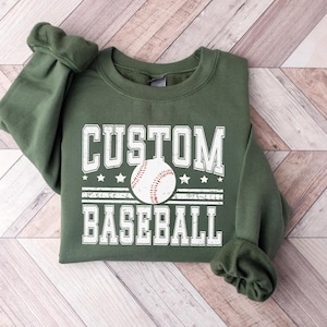May include: An olive green sweatshirt with the words "CUSTOM BASEBALL" in white block letters. A baseball graphic is centered between the words. The sweatshirt has a crew neck and long sleeves.