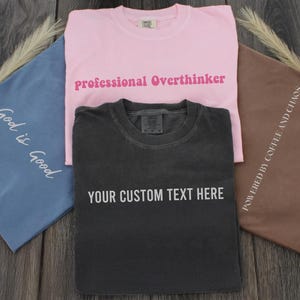 three t-shirts with custom text printed on them, laid out on a wooden surface.