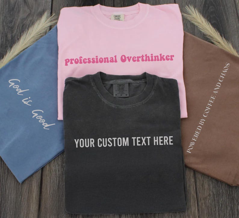 three t-shirts with custom text printed on them, laid out on a wooden surface.