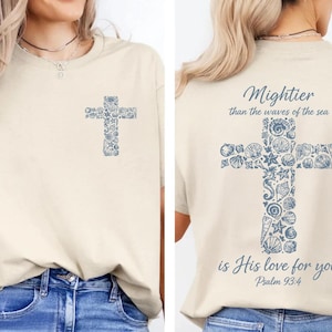 May include: Cream-colored t-shirt with a blue cross design on the front and back. The cross is made of seashell and floral patterns. The back of the shirt has the text "Mightier than the waves of the sea is His love for you Psalm 93:4."