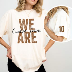 May include: A white t-shirt with the text "We Are Custom Team" in brown distressed lettering. The shirt also has a brown number 10 on the sleeve.