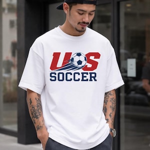 A man wearing a white t-shirt with the text "U.S. SOCCER" printed on it, standing in front of a building.