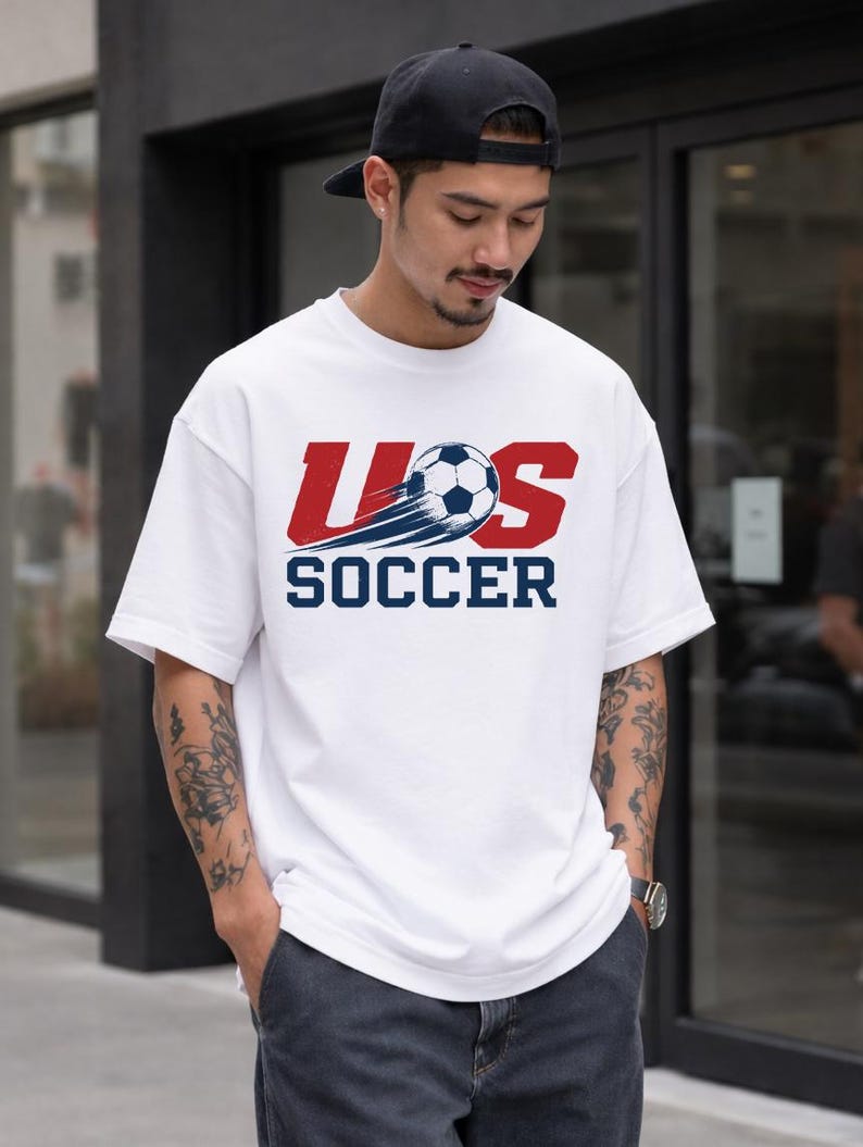 A man wearing a white t-shirt with the text "U.S. SOCCER" printed on it, standing in front of a building.