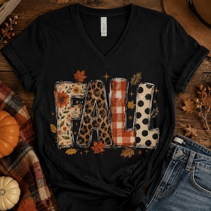 May include: A black V-neck t-shirt featuring the word FALL in large block letters. Each letter has a distinct pattern: floral with orange flowers, leopard print, red and white buffalo plaid, and black polka dots on a cream background.