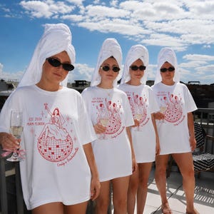 May include: A white t-shirt featuring a red graphic of a person sitting on a disco ball, holding a glass, with the text EST 2026 MIAMI FLORIDA and Emily's Bachelorette. The shirt is worn with a white towel wrapped around the head and black sunglasses.