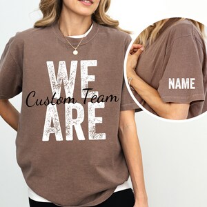 May include: A brown t-shirt with the words "We Are Custom Team" printed in white on the front. The back of the shirt has a blank space for a name.