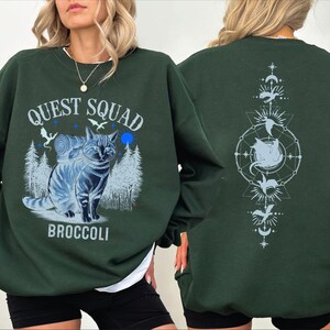 May include: A dark green sweatshirt with a graphic print of a cat with a backpack and the text "Quest Squad Broccoli" on the front. The back of the sweatshirt has a graphic print of a cat in a circle with stars and moons.