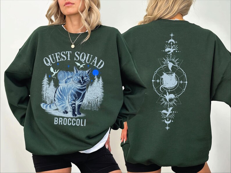 May include: A dark green sweatshirt with a graphic print of a cat with a backpack and the text "Quest Squad Broccoli" on the front. The back of the sweatshirt has a graphic print of a cat in a circle with stars and moons.