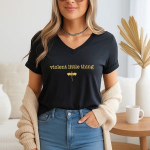 A woman wearing a black t-shirt with the text "violent little thing" and blue jeans, standing in a room with a beige sweater and a coffee mug on a table.
