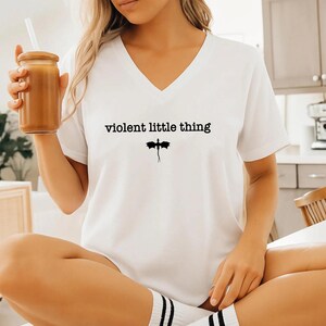 A woman wearing a white t-shirt with the text "violent little thing" printed on it, sitting in a kitchen and holding a drink.