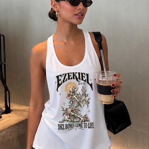 May include: A white sleeveless tank top featuring a graphic print of a skeleton kneeling among flowers with a sunburst background. The text reads EZEKIEL 37:1-10 and DRY BONES COME TO LIFE in a dark, stylized font. The design includes small star accents.