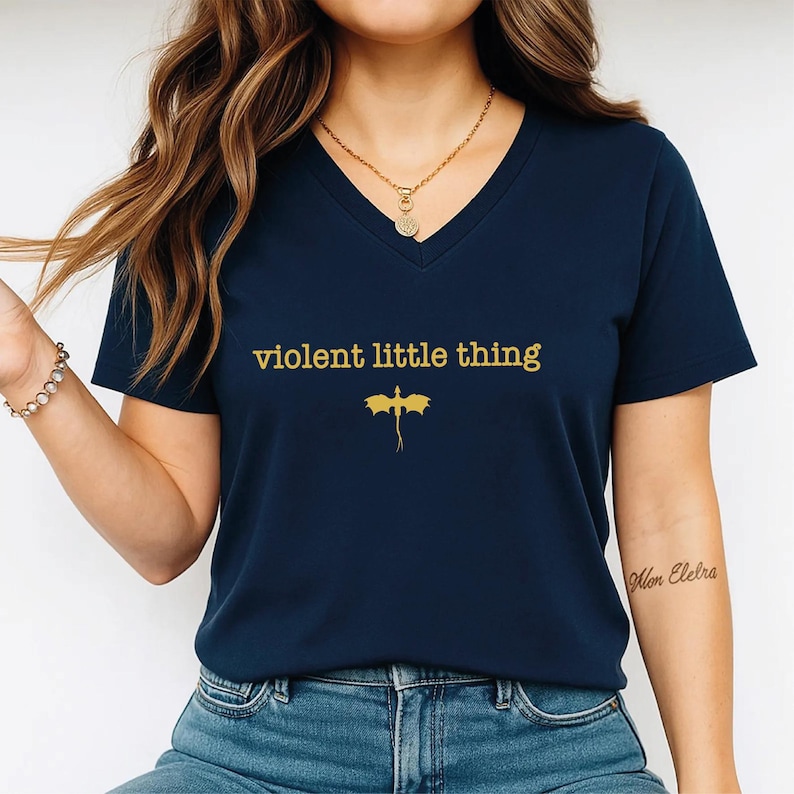 A woman wearing a navy blue t-shirt with the text "violent little thing" printed on it, along with a dragon graphic. She is also wearing a necklace and has a tattoo on her arm.