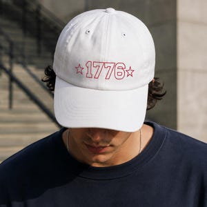 May include: White baseball cap with the red text "1776" and star accents. The hat is made of a soft material and has a curved brim. The cap is a classic style, suitable for casual wear.
