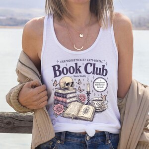 A woman wearing a white tank top with a graphic design of books, a skull, and flowers, standing near a body of water.