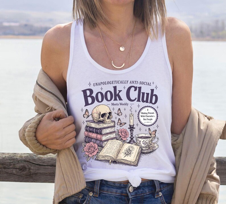 A woman wearing a white tank top with a graphic design of books, a skull, and flowers, standing near a body of water.