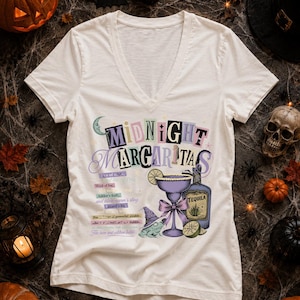 May include: A cream-colored V-neck t-shirt featuring the text Midnight Margaritas, a purple margarita glass, a tequila bottle, a frog wearing a witch hat, and a list of ingredients including eye of newt and wool of bat in colorful text.