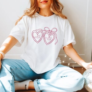 May include: White t-shirt featuring a magenta heart-shaped glasses design with the text "Custom Text" inside each lens. The glasses are connected by a bow. The shirt is made of a soft material.