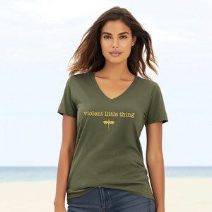 A woman wearing a green t-shirt with the text "violent little thing" printed on it, standing on a beach with the ocean in the background.
