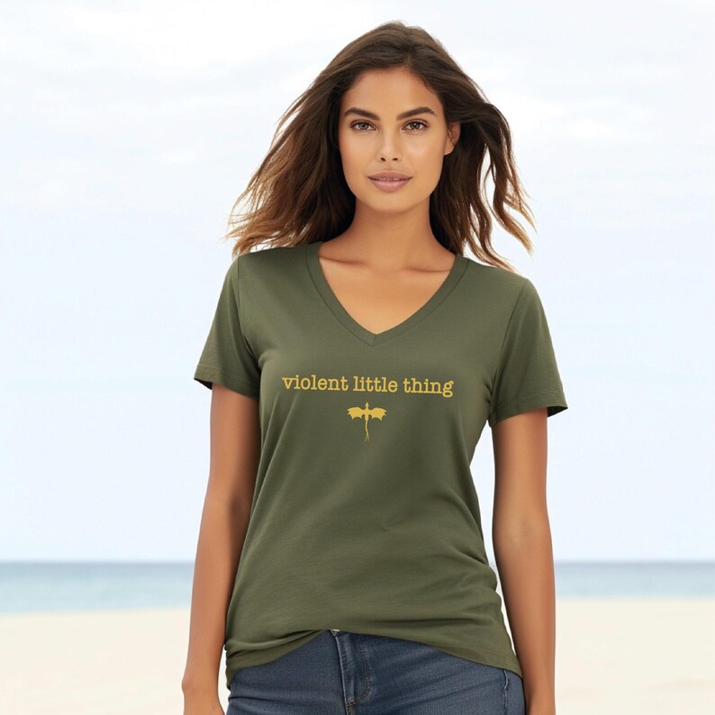 A woman wearing a green t-shirt with the text "violent little thing" printed on it, standing on a beach with the ocean in the background.