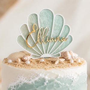 May include: A cake topper shaped like a seashell with a white outline and a textured sage green interior. The name Elliana is written in gold cursive script across the center of the shell. The topper is placed on a cake decorated with sand and shells.