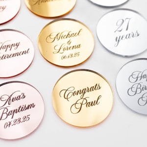 May include: Assorted metallic round tokens with engraved text for various occasions. Gold, silver, and rose gold tokens feature phrases like "Happy Retirement," "Michael & Lorena," and "Happy Birthday Robin."