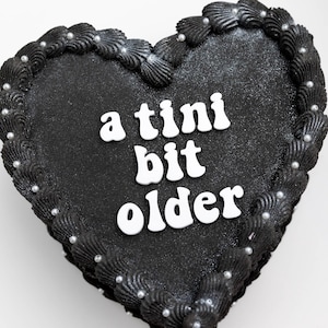 May include: A heart-shaped cake with black frosting and the words "a tini bit older" in white. The cake is decorated with black frosting swirls around the edge and small silver sprinkles.