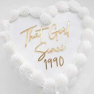 May include: A heart-shaped cake with white frosting piped in a shell border around the edge. Centered on the smooth white surface is a gold-colored cake topper with the text That Girl Since 1990 written in a cursive and print font combination.