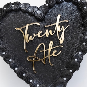 May include: A black heart-shaped cake with a gold glitter finish and a gold inscription that reads "Twenty Ate". The cake is decorated with black frosting and white pearl sprinkles.